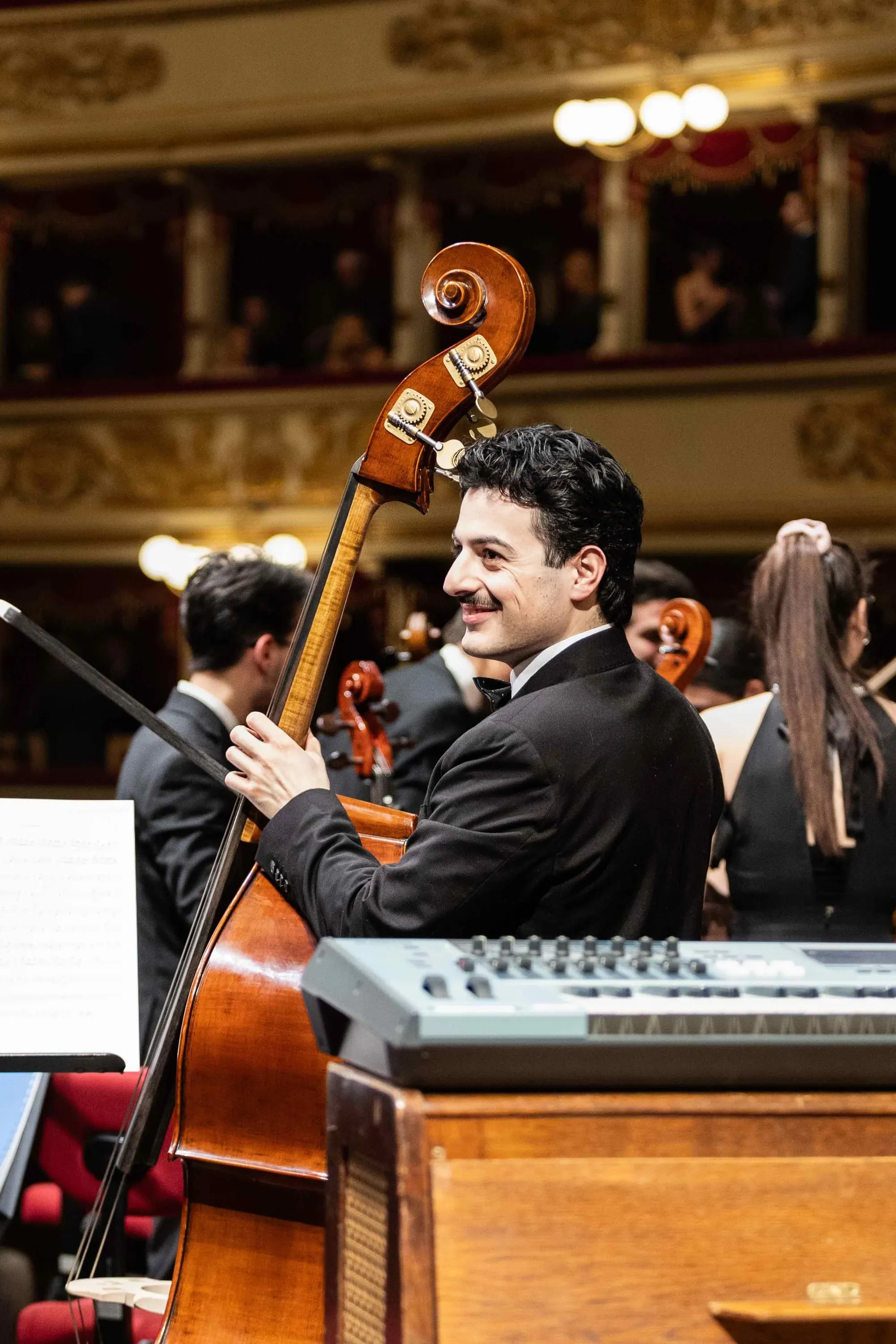 Un contrabbassista sorride tra i musicisti, con i palchi della Scala